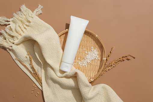 Close-up of an unbranded cosmetic tube and white rice on a flat winnowing basket on a brown background. Cosmetics contain natural ingredients. Top view.