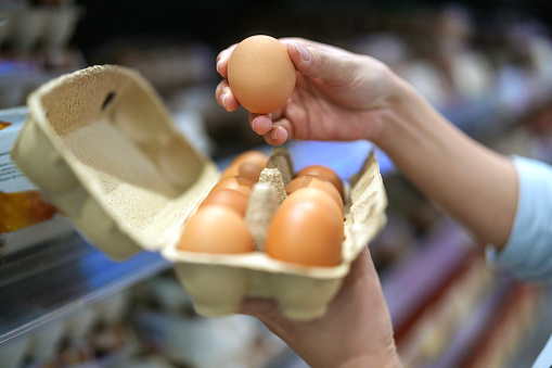 In this cropped image, a customer's hands are inspecting a box of eggs in a grocery store, emphasizing the careful scrutiny and selection process while shopping for fresh produce.
