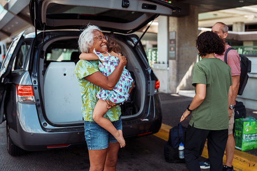 A beautiful senior woman of Hawaiian and Chinese descent who just arrived in Hawaii with her Caucasian husband to visit family, smiles with excitement while affectionately greeting her three year old granddaughter outside the airport.