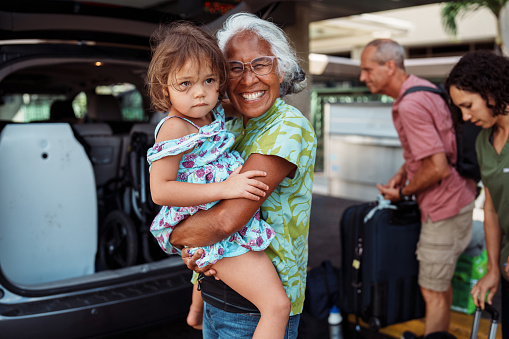 A beautiful senior woman of Hawaiian and Chinese descent who just arrived in Hawaii with her Caucasian husband to visit family, smiles with excitement while affectionately greeting her three year old granddaughter outside the airport.