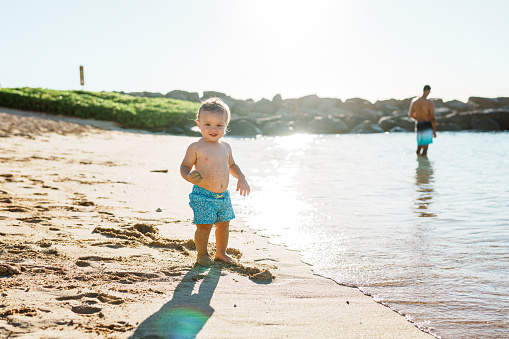 A multiracial one year old boy wearing a swimsuit walks by himself along the ocean shoreline during a beautiful tropical beach day.