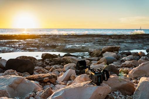 A large camera belonging to an unknown photographer sits on a rock on the shoreline of an ocean during sunset.