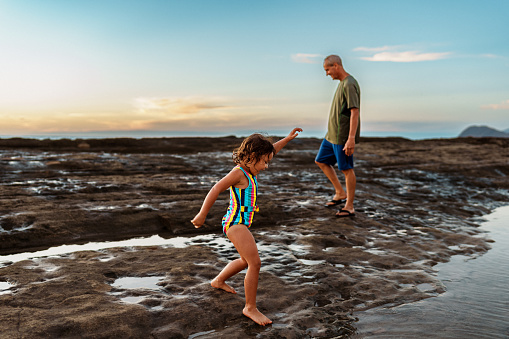 An active and healthy senior man of Caucasian descent has fun exploring and walking along the beach at sunset with his cute three year old Eurasian granddaughter while on vacation in Hawaii.