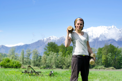 Happy farmer woman with firewood on a green field with grass and trees and snow-capped mountains in switzerland