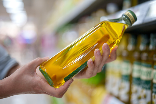In a close-up shot, a woman's hands are reading the label on a bottle of cooking oil taken from the shelf in a grocery store, ensuring product freshness and safety.