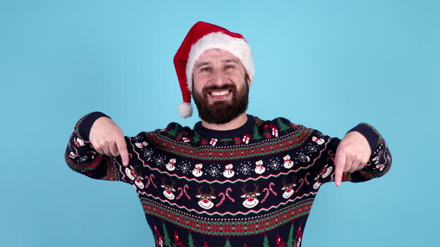 Shocked bearded man in new year clothes pointing with hands down on blue isolated background, surprised guy in santa hat showing and advertising copy space.