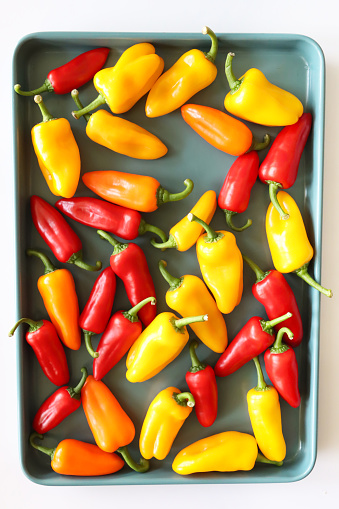 Stock photo showing close-up, elevated view of cooking tray covered in mini red, orange and yellow bell peppers on white background.