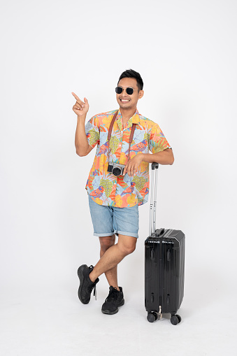 A fun, cheerful Asian man tourist in summer clothes is pointing his finger up at an empty space on isolated white studio background. recommend, showing option, advice gesture