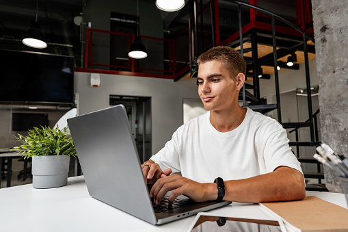 Casual young man in white t-shirt sitting at white desk looking at laptop screen at modern office close up