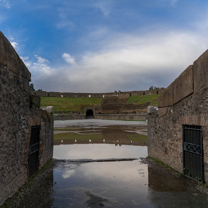 Pompei, Italy - 25 November, 2023: view of the amphitheatre and entrance with reflections in a puddle in the ancient city of Pompeii