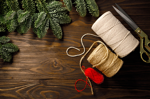 scissors, cream coloured and red twisted cotton ropes and burlap cord on dark wooden table decorated with spruce branches. Concept of handmade crafting, Christmas and winter leisure. Top view