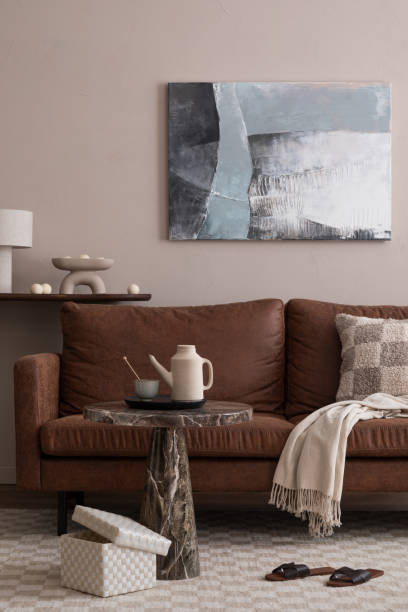 istock Interior design of cozy living room interior with mock up poster frame, brown sofa, patterned pillow, plaid, slippers, round coffee table, beige pitcher and personal accessories. Home decor. Template. 1869299183