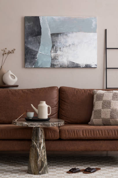 istock Interior design of cozy living room interior with mock up poster frame, brown sofa, patterned pillow, plaid, slippers, round coffee table, beige pitcher and personal accessories. Home decor. Template. 1869300221