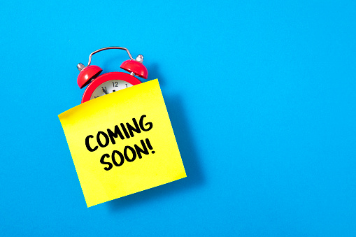 Coming soon message on adhesive note with red alarm clock on blue background