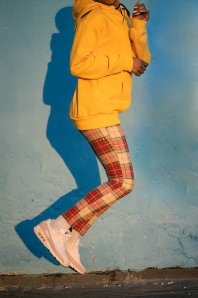 An unrecognisable woman wearing plaid trousers, trainers and a yellow sweatshirt is jumping with her knees bent and feet in the air against a blue background