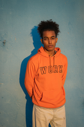 A chilled looking Afro-Caribbean man standing against a blue background, wearing an orange sweatshirt with his hands behind his back as he looks seriously at the camera