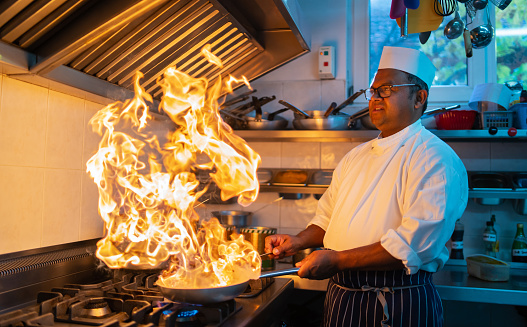 An Indian chef is looking at the sea of flames as he is cooking on an industrial stove in a restaurant kitchen