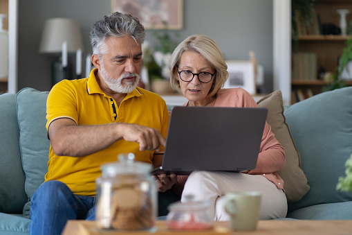 A couple of silver surfers are using a laptop, sitting together on a sofa in a living room