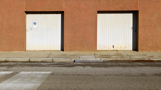 Old white garage doors on brown building facade at the roadside. Concrete sidewalk and city street in front. Background for copy space.