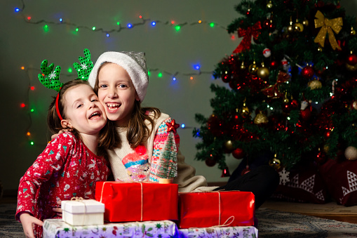Portrait of two happy girls sitting on the floor in front of Christmas tree during the night