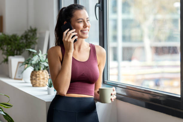 Sporty woman using mobile phone while drinking a cup of coffee after training at home.