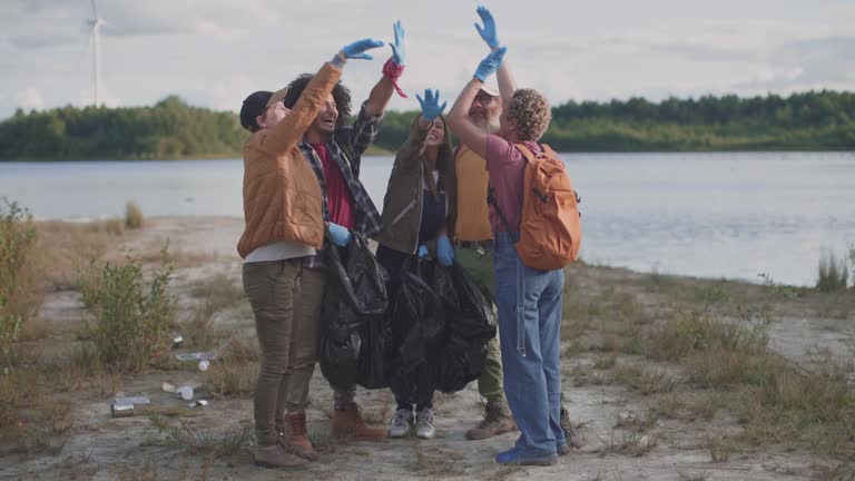 Volunteer team giving high five in nature environment cleaning riverbank from rubbish with plastic bags and gloves