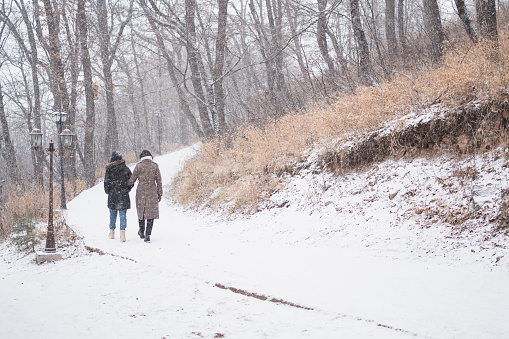 Two women walking by snowy trail in winter urban park. full body from behind
