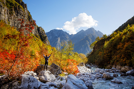 A happy woman with raised arms is standing on a rock next to a mountain stream. High mountains visible in the distance. Shot close to Theth in northern Albania.