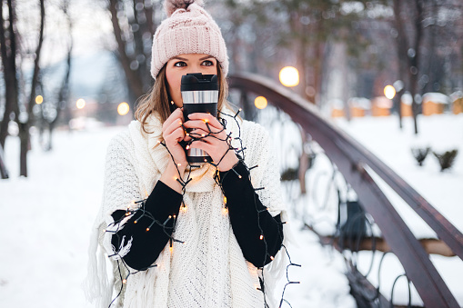 Beautiful young woman holding a cup of coffee and Christmas lights in a snowy park in winter.