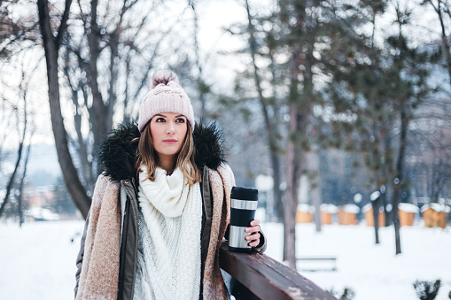Beautiful young woman drinking coffee in a snowy park in winter.