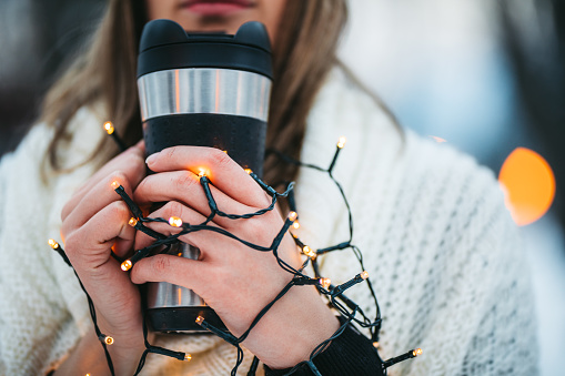 Beautiful young woman holding a cup of coffee and Christmas lights in a snowy park in winter. Close-up.