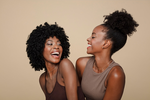 Two excited young women wearing brown clothes standing against beige background, embracing and smiling.