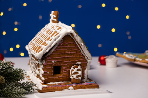 Stock photo showing close-up view of a snowy clearing, conifer forest scene. Three gingerbread men skating in front of homemade, gingerbread house decorated with white royal icing surrounded by model fir trees on white, icing sugar snow against a snowy blue background.