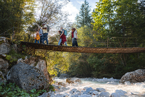 A multiracial family wearing casual clothes are walking over a hanging bridge over a river outside at the nature. The landscape at the back is full of trees and beautiful.