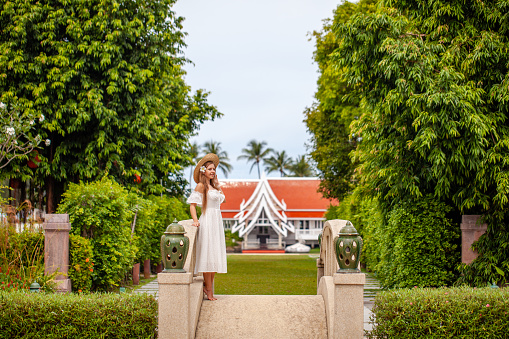 Elegant woman in white dress enjoying leisurely walk in tropical garden. Travel and relaxation.