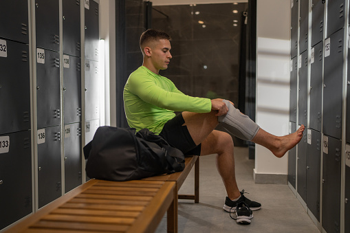 Fit man putting on a kneepad before training, he is in gym locker room.