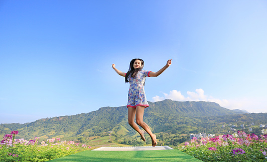 Jumping Young Asian woman feeling free with arms wide open at beautiful trees and mountains on blue sky.