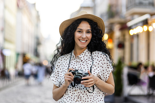 Portrait of young beautiful woman tourist with camera, Hispanic woman with curly hair in hat walking in evening city smiling and looking at camera close up.