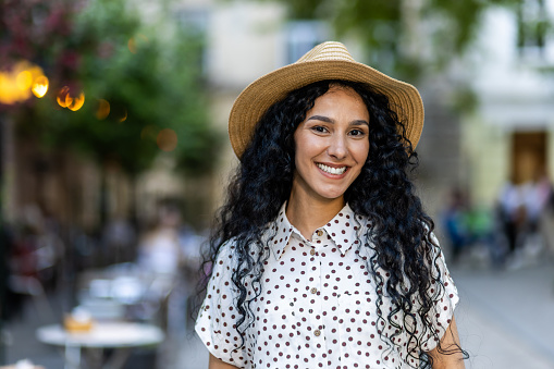 Beautiful young Latin American woman portrait, woman walking in evening city in hat with curly hair in warm weather, smiling and looking at camera close up.