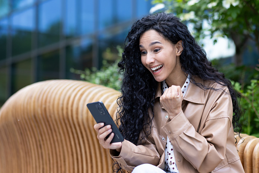 Young happy woman celebrating victory triumph success, satisfied hispanic business woman holding phone received online win notification, sitting on bench outside office building.