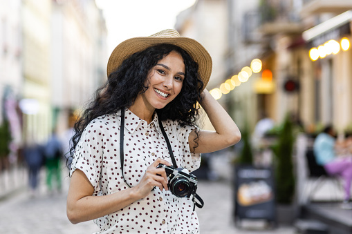 Portrait of young beautiful woman tourist with camera, Hispanic woman with curly hair in hat walking in evening city smiling and looking at camera close up.