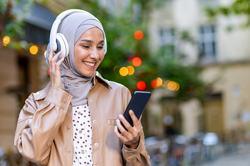 Young beautiful woman walking in the city, Muslim woman in hijab on evening walk in headphones listening to music smiling joyfully using smartphone app.