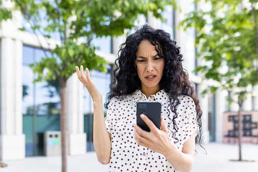 Sad disappointed woman received online notification with bad news on her phone, businesswoman walking outside office building, using application on smartphone, reading social media unsatisfied.