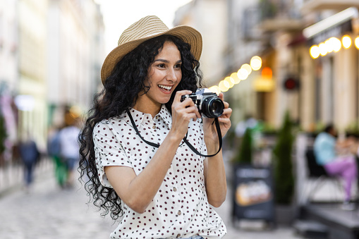 Young beautiful hispanic woman with curly hair walking in the evening city with a camera, female tourist on a trip exploring historical landmarks in the city.