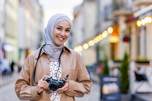 Portrait of young beautiful Muslim woman in hijab, tourist walking in evening city with camera, woman traveling to different countries