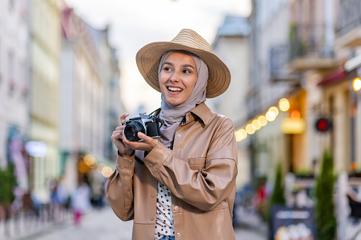 Young beautiful woman walking in the evening city in hijab, tourist with camera and wearing a hat inspects the historical city smiling with satisfaction, Muslim woman on a trip.