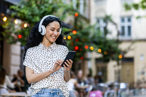 Young beautiful hispanic woman walks evening city in headphones, student listens to music online, uses an application on a smartphone, holds the phone in her hands, reads messages, smiles.