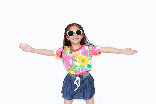 Happy little Asian kid girl stretch arms wide open wearing a floral pattern summer dress and sunglasses isolated on white background. Summer and fashion concept.
