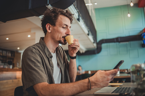 A young hipster man in a cafe drinking coffee and using a mobile phone to surf the Internet or correspond on social networks