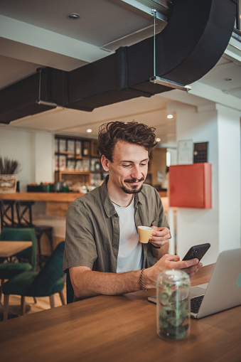 A young hipster man in a cafe drinking coffee and using a mobile phone to surf the Internet or correspond on social networks